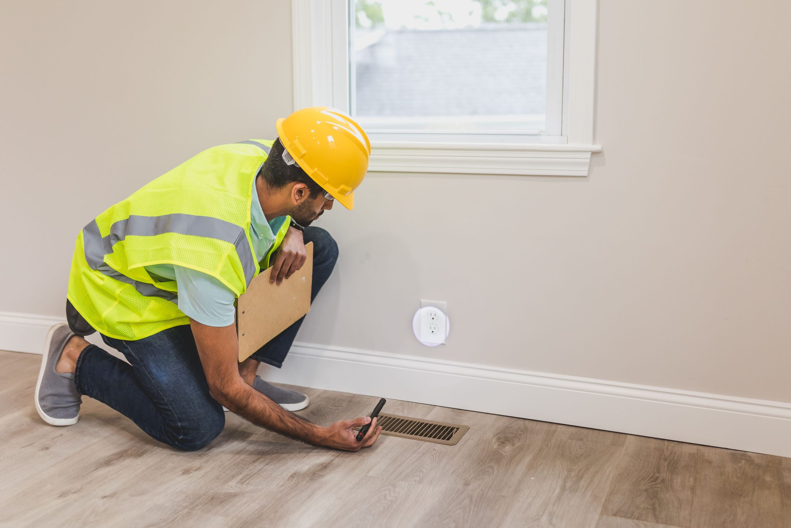 Home inspector in a hard hat checking a floor vent with a clipboard during a home insurance inspection